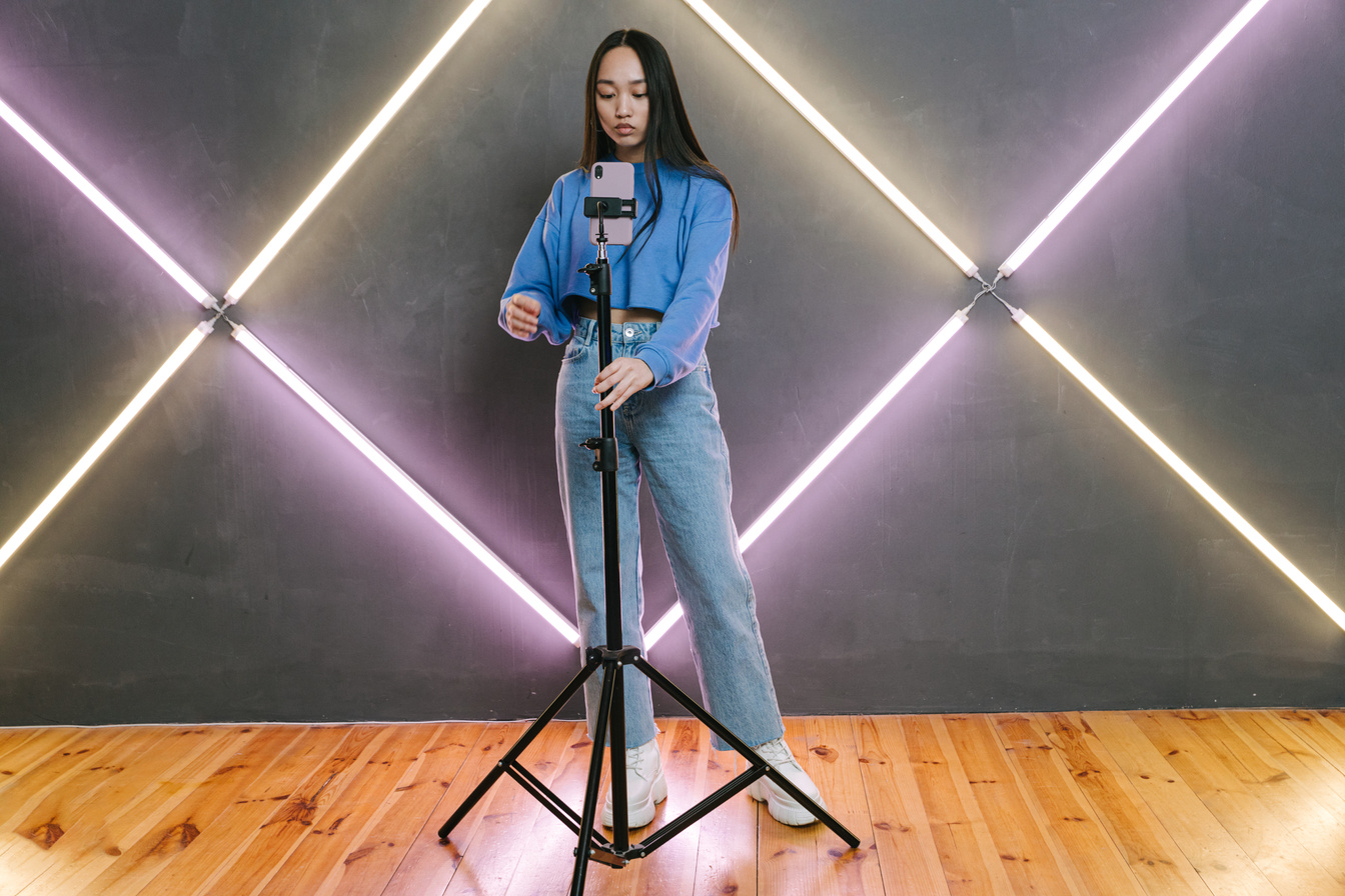 A Woman in Blue Sweater and Denim Jeans Holding a Tripod with Mobile Phone